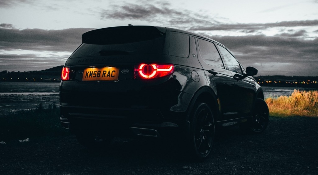 "Achterkant van een zwarte Land Rover Discovery Sport met brandende achterlichten in de schemering, geparkeerd bij water.